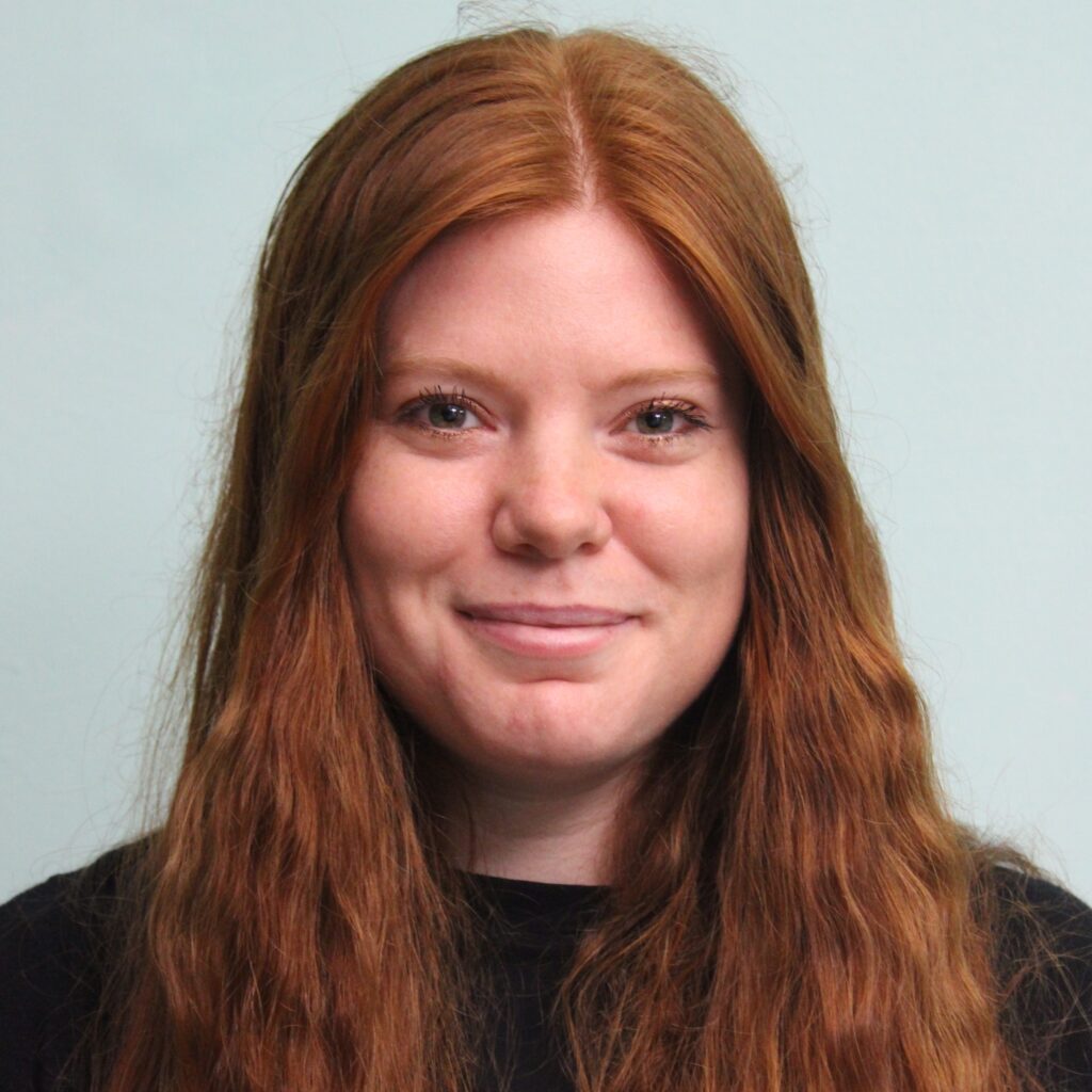 Porträt frontal: Jenny hat lange rote Haare, die sie offen trägt. Sie hat grüne Augen und lächelt in die Kamerat