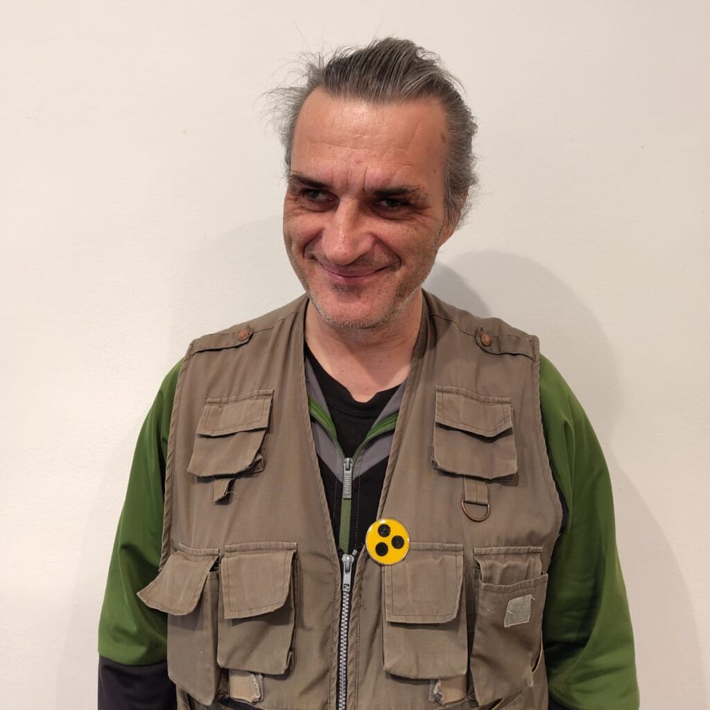 Man sieht den Trainer von vorne. Er lächelt in die Kamera. Über einem grünen Pulli trägt er eine Weste, auf der das Blindenabzeichen ist.