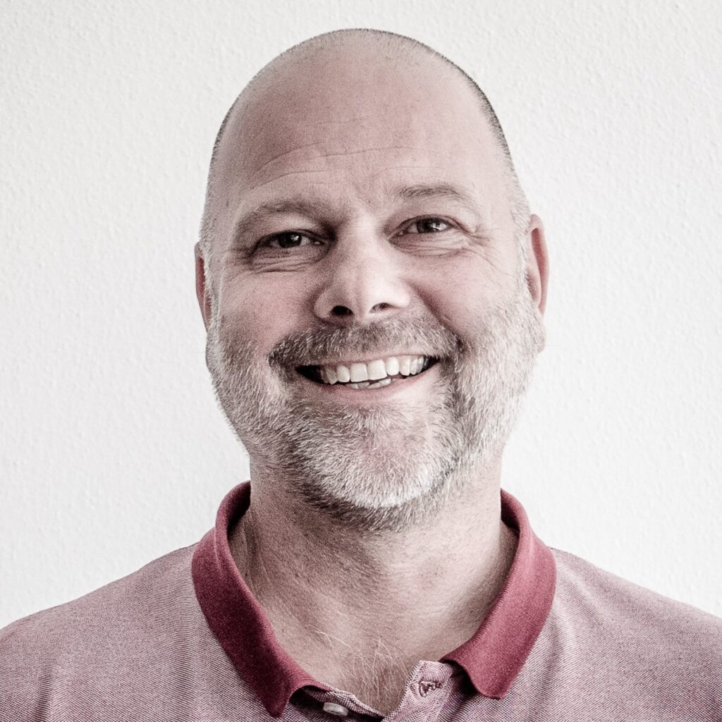 Andi Ausserhofer lächelt in die Kamera. Er hat wenig Haare am Kopf und einen schon weißen Bart. Er trägt ein rotes T-Shirt.