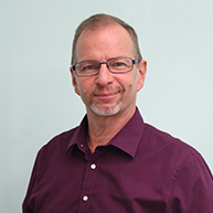 Manfred Philippich hat einen Bart mit schon etwas weiß darin und eine Brille. Er trägt ein rotes Hemd.