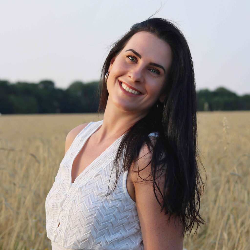 Veronika steht auf einer Wise und trägt ein weißes Kleid. Sie hat lange schwarze Haare und lächelt in die Kamera.