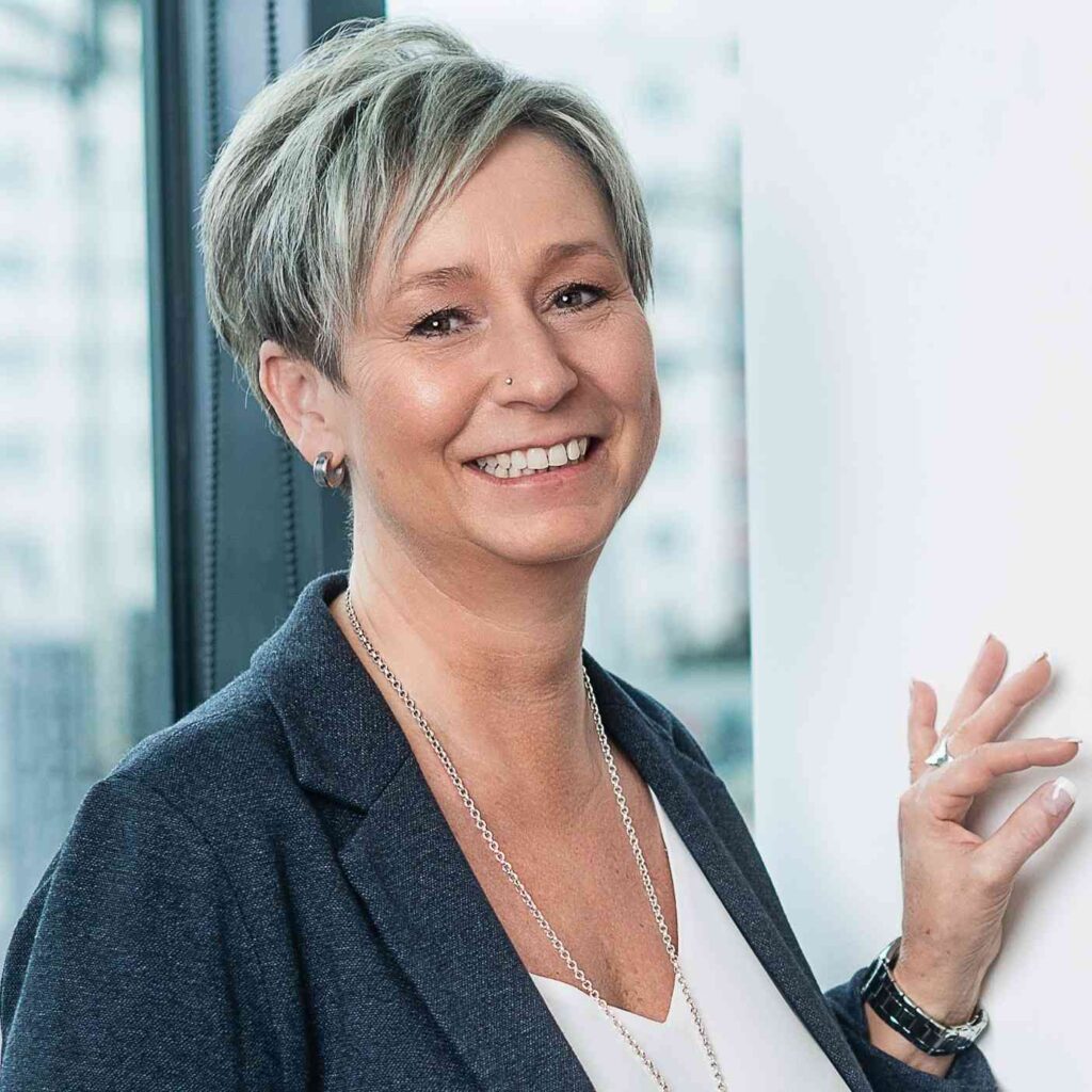 Sandra Moosbacher hat blone kurze Haare und keine Brille. Sie trägt einen blauen Blazer, eine weißes T-Shirt und eine Kette.