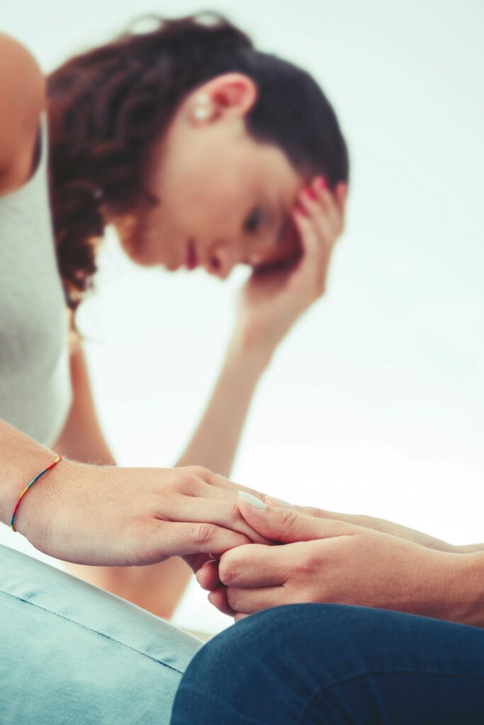 Vordergrund: zwei Personen halten sich an den Händen; Hintergrund: eine Person mit gesenktem Kopf stützt die Stirn mit der Hand.