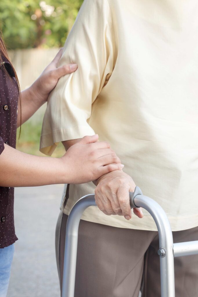 Eine Person stützt und begleitet eine ältere Person mit Rollator, Hand liegt unterstützend am Arm.